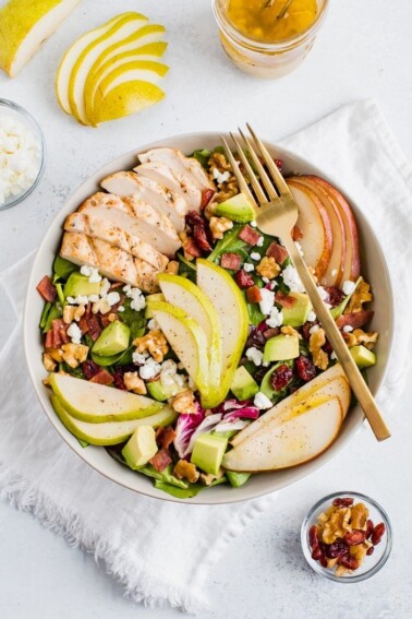 A full bowl of a salad topped with pears, grilled chicken, turkey bacon, cranberries, goat cheese, walnuts, and avocado with a fork.