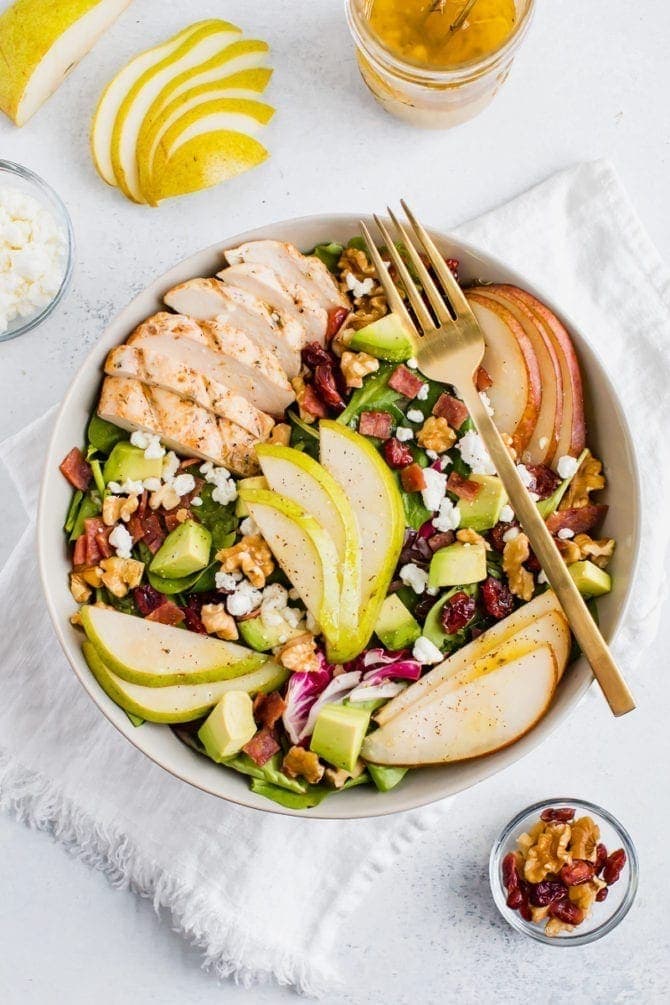 A full bowl of a salad topped with pears, grilled chicken, turkey bacon, cranberries, goat cheese, walnuts, and avocado with a fork.