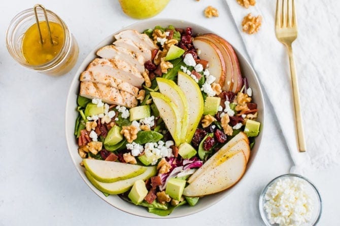 A salad topped with pears, grilled chicken, turkey bacon, cranberries, goat cheese, walnuts, and avocado.