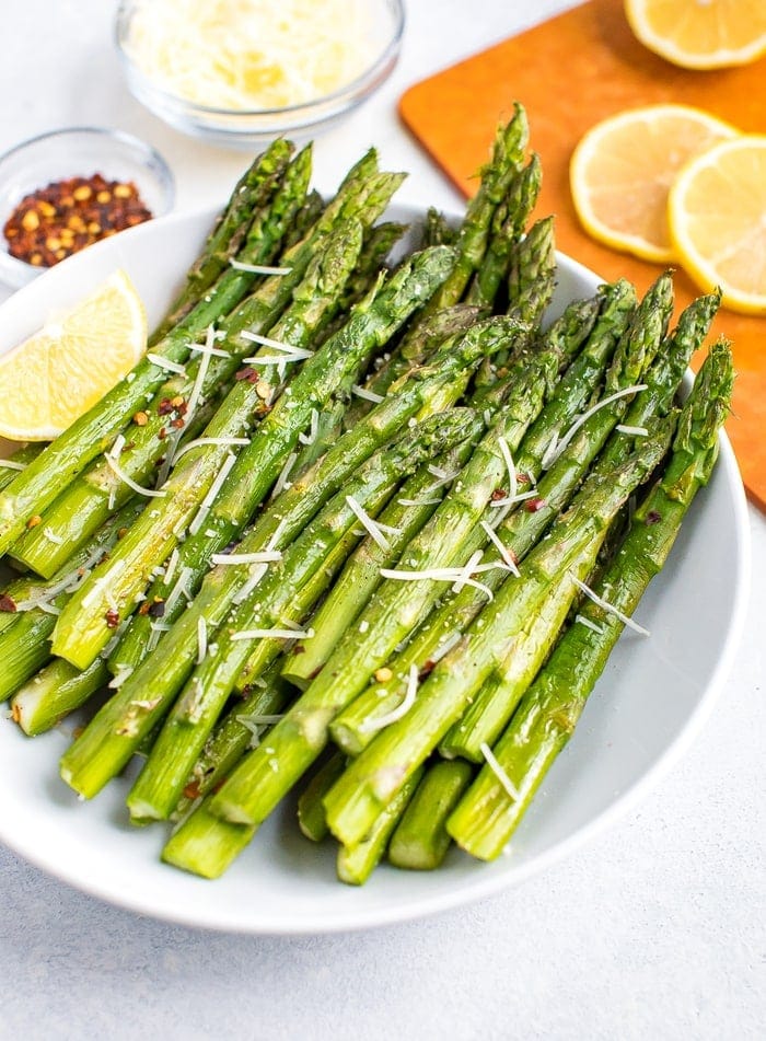 Plate of roasted asparagus topped with parmesan, red pepper flakes, and a lemon wedge on the side. Beside the bowl are smaller bowls filled with red pepper, parmesan, and a cutting board with lemon slices.