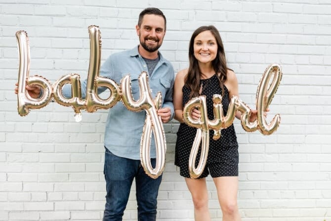 Man and woman holding baby girl balloons against a white brick wall.