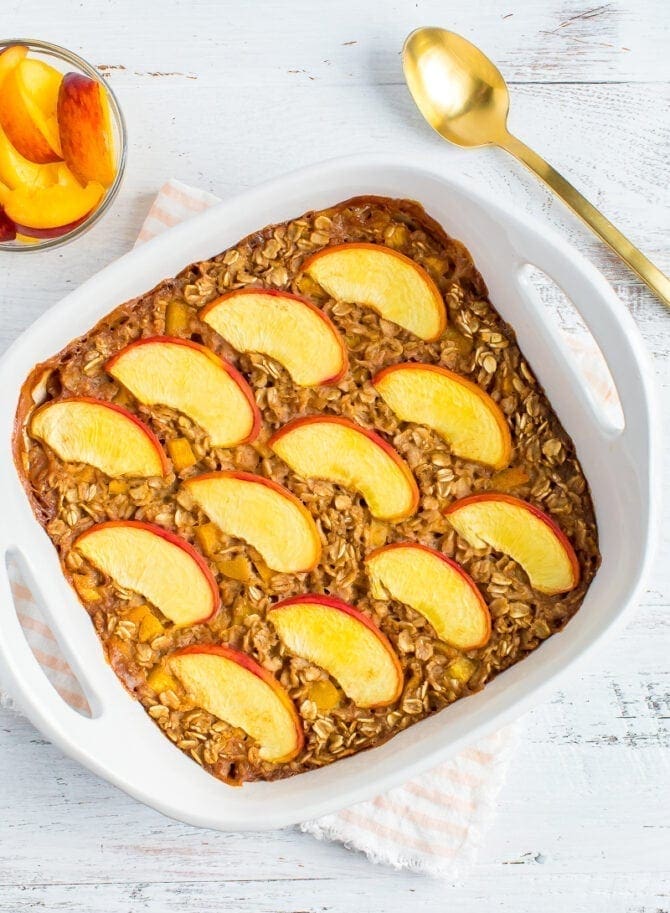 Baked oatmeal in a square dish and topped with peaches. A bowl of peach slices and a spoon are to the side.