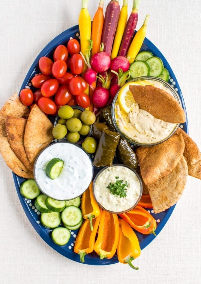 A vegetarian mezze platter with carrots, radishes, tomatoes, cucumber slices, green olives, domades, chips, peppers, hummus and dips.