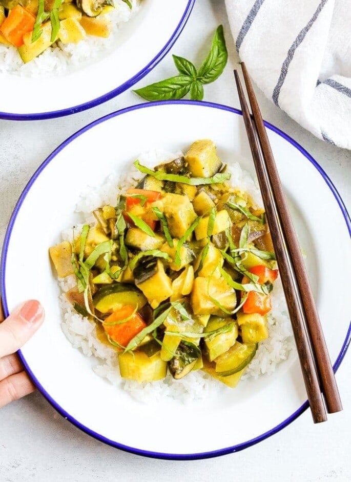 Hand holding a plate of summer vegetable coconut curry served over rice and topped with basil. Two chop sticks are on the side of the bowl.