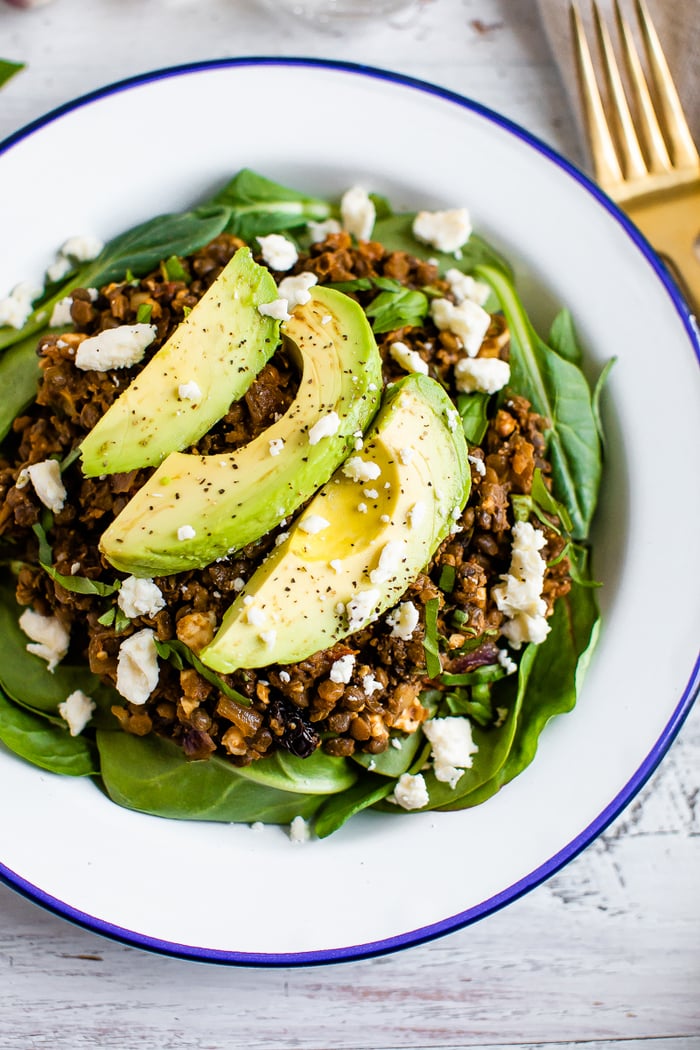 A plate with a bed of fresh spinach topped with a warm Mediterranean salad, topped with feta cheese and sliced avocado.