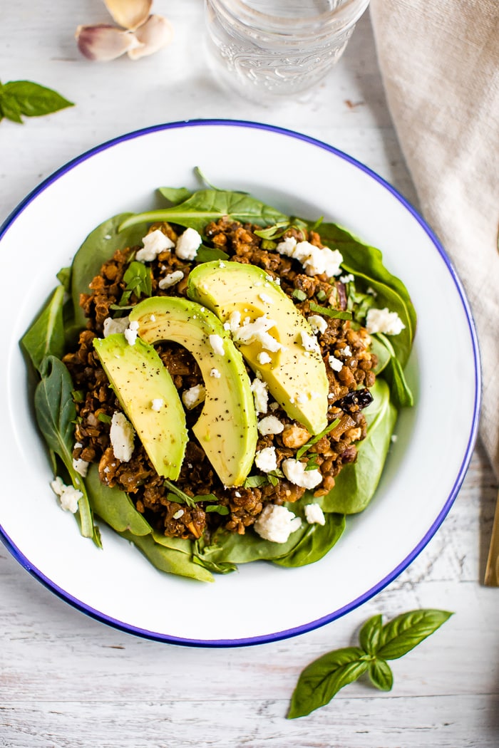A serving of fresh spinach topped with a warm Mediterranean salad, topped with feta and avocado.