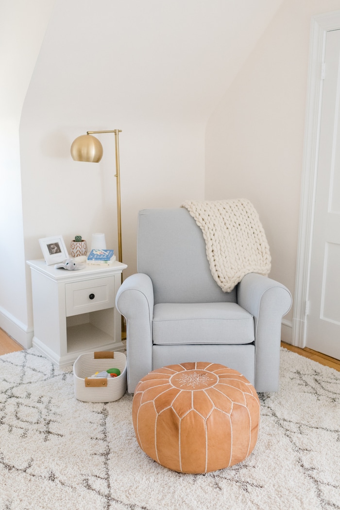 Baby girl nursery with a blue gray glider, gold flour lamp and leather pouf.