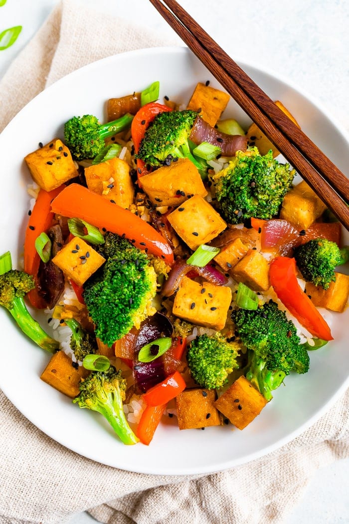 Bowl of rice with tofu and vegetable stir fry on top. Chopsticks are resting on the bowl.