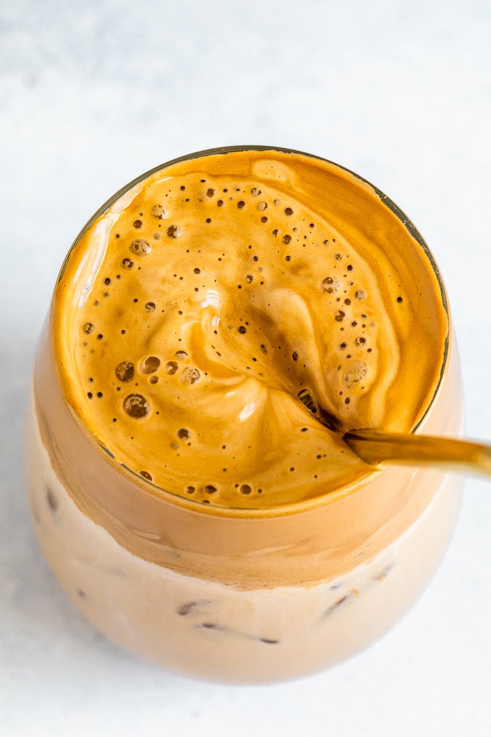 Foamy and bubbly coffee foam in a glass over iced dairy-free milk. Gold straw is in the glass.