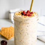 A serving of cherry cheesecake overnight oats in a mason jar topped with dried cherries and graham crackers in the background.