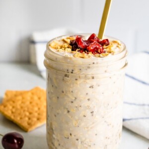 A serving of cherry cheesecake overnight oats in a mason jar topped with dried cherries and graham crackers in the background.