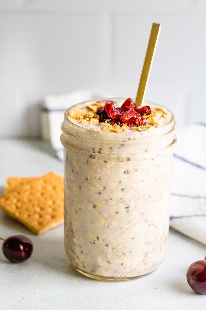 A serving of cherry cheesecake overnight oats in a mason jar topped with dried cherries and graham crackers in the background.