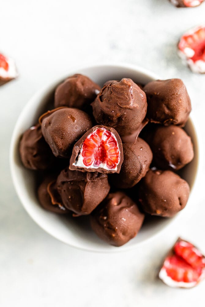 Bowl of chocolate covered frozen raspberries with yogurt. One raspberry is cut in half. Raspberries scattered on the table around the bowl.