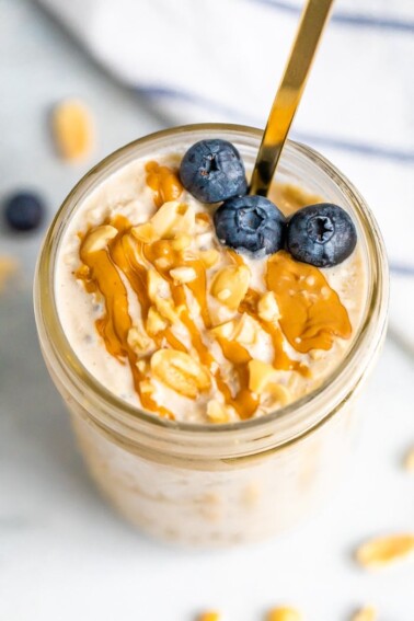 A serving of peanut butter overnight oats in a mason jar topped with fresh blueberries, chopped peanuts and a peanut butter.
