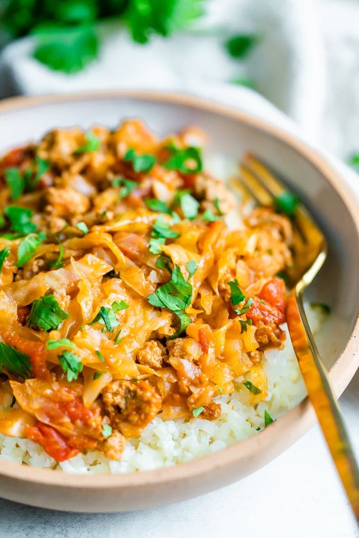 Close up photo of unstuffed cabbage roll bowl. Cabbage and turkey are mixed with a tomato sauce and served over rice. Gold fork is resting inside white ceramic bowl.