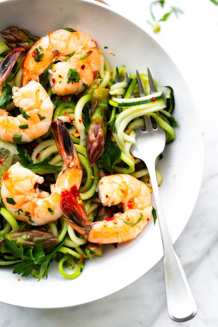 A bowl served with zucchini noodles, asparagus and shrimp garnished with red pepper flakes.