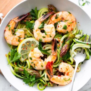 A bowl with zucchini noodles, asparagus and shrimp garnished with lemon and red pepper flakes.