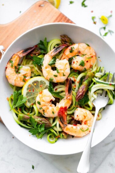 A bowl with zucchini noodles, asparagus and shrimp garnished with lemon and red pepper flakes.