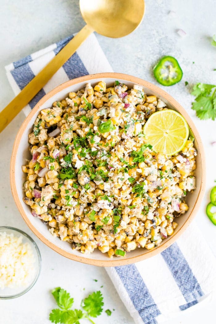 Bird's eye view photo of a bowl of Mexican Street Corn Salad topped with lime and fresh cilantro. Bowl is resting on a striped napkin and next to a gold spoon and bowl of cheese.