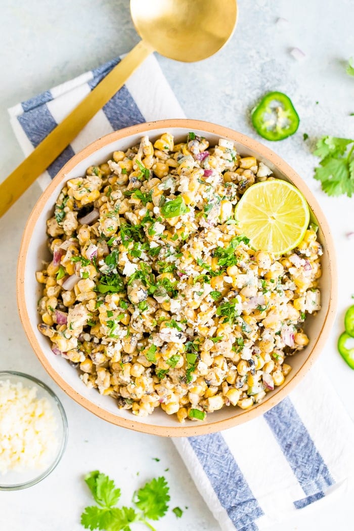 Bird's eye view photo of a bowl of Mexican Street Corn Salad topped with lime and fresh cilantro. Bowl is resting on a striped napkin and next to a gold spoon and bowl of cheese.