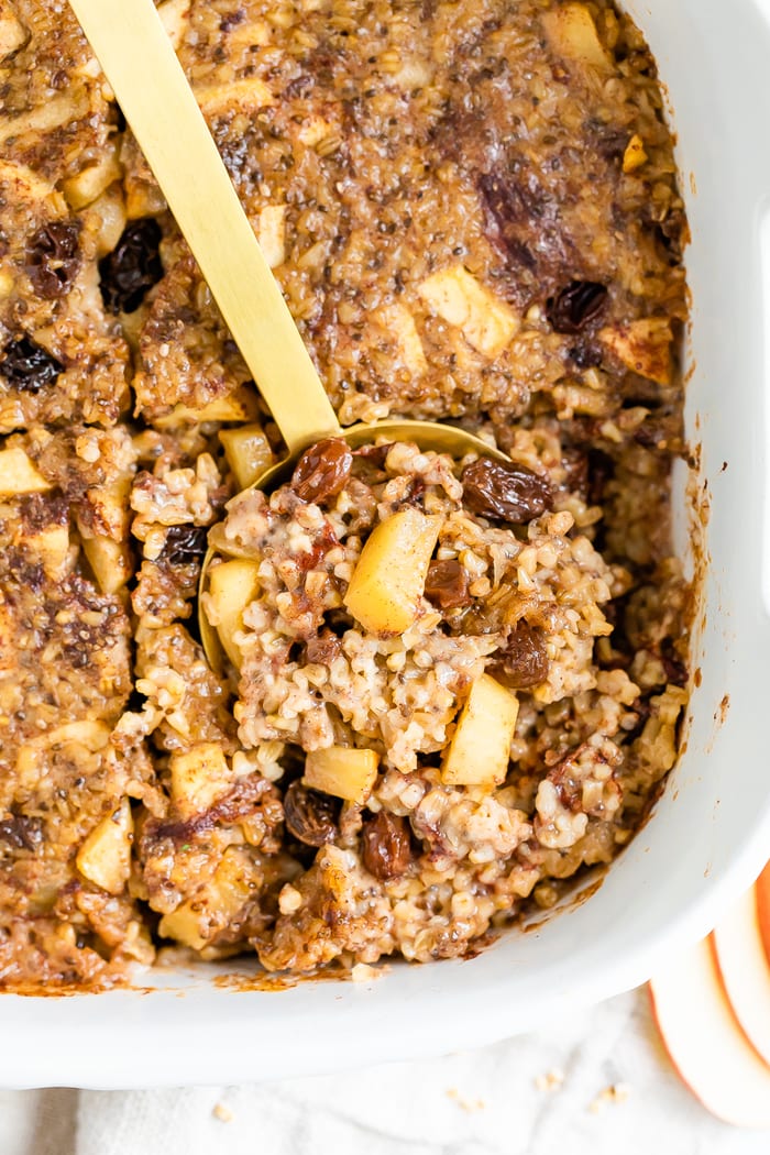 Overhead photo of apple cinnamon baked steel cut oatmeal in a white baking dish with a gold spoon scooping out a portion.