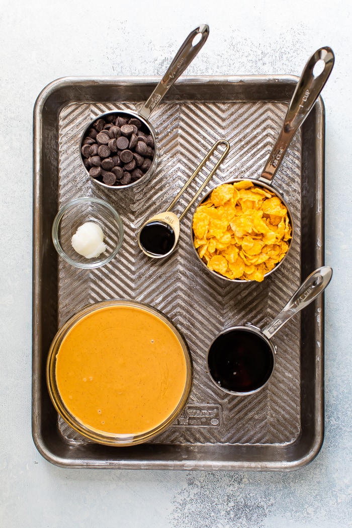 Overhead photo of baking sheet with ingredients measured out. Peanut butter, maple syrup, coconut oil, corn flakes and chocolate chips in measuring cups.