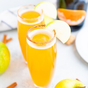 Two tall glasses of mimosas with a cinnamon sugar rim and an apple slice for garnish.