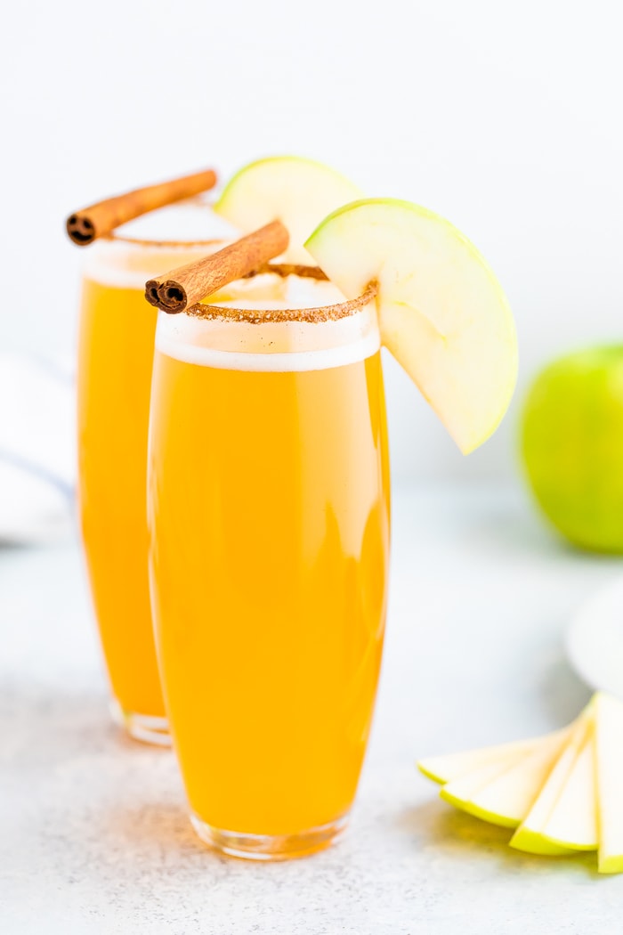 Two tall glasses of apple mimosas with a cinnamon sugar rim, cinnamon sticks and a green apple slice for garnish.