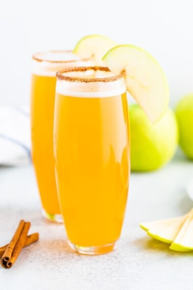 Two tall glasses of mimosas with a cinnamon sugar rim and an apple slice.