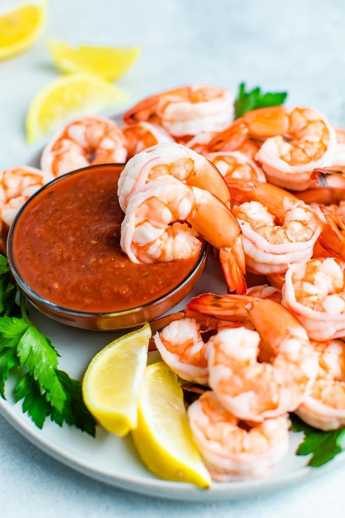 Poached shrimp on a plate with cocktail sauce and garnished with lemon and parsley.