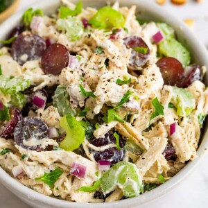 A large bowl of shredded chicken, celery, red onion, red grapes, herbs, and spices.