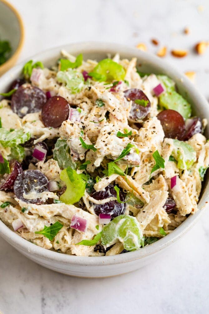 A large bowl of shredded chicken, celery, red onion, red grapes, herbs, and spices.