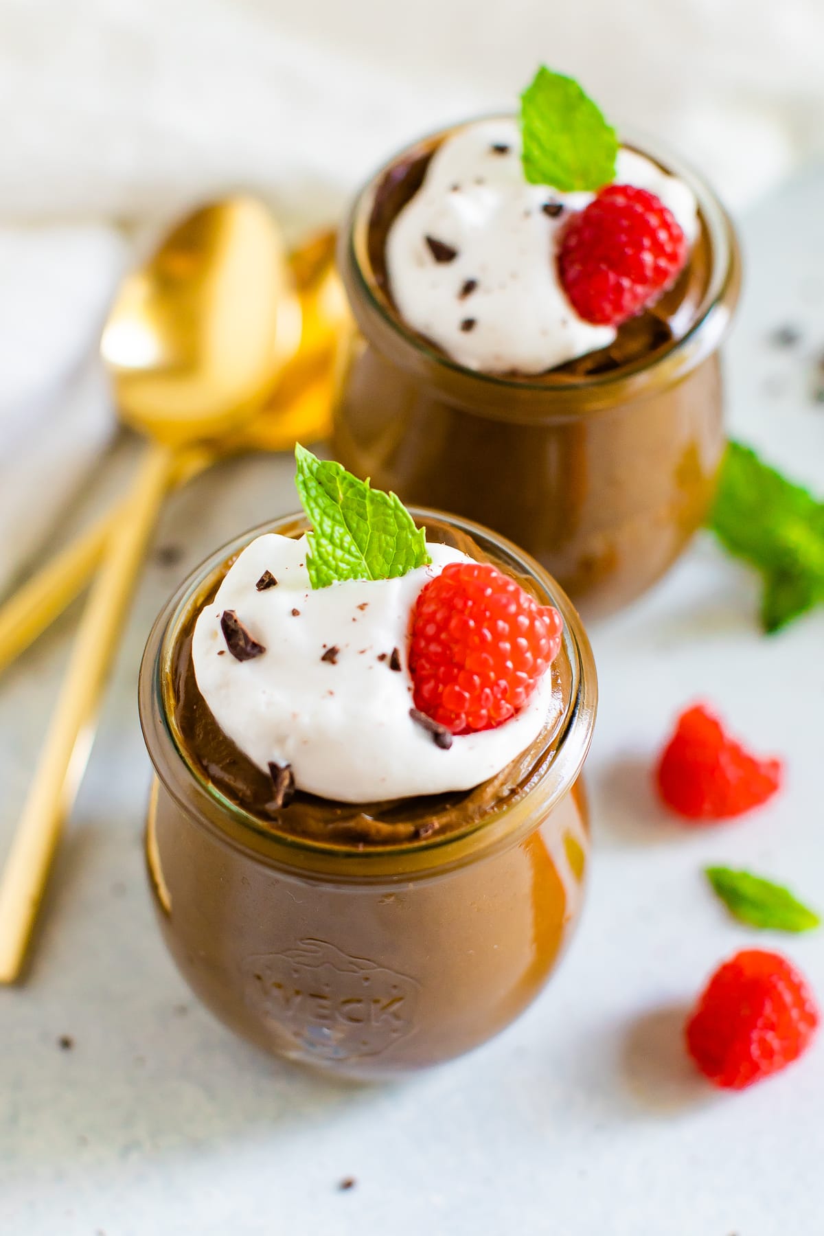 Two jars of avocado chocolate pudding topped with whipped cream, raspberries chocolate shavings and a mint leaf.