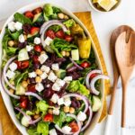 A large oval plate with a fresh Greek salad with cucumber, tomatoes, feta, olives, and lettuce.