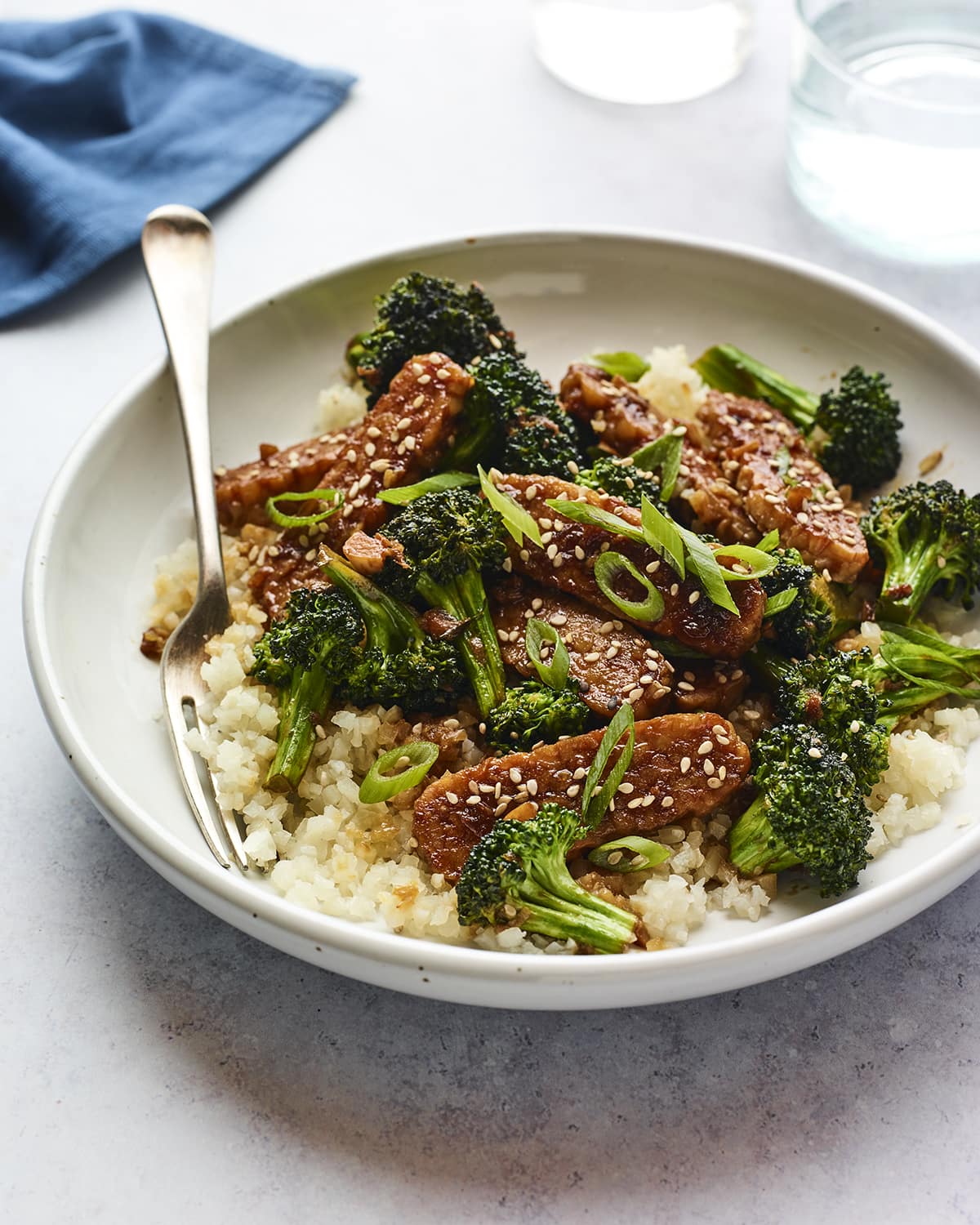 A serving of teriyaki tempeh and broccoli over white rice in a shallow bowl with a metal fork.