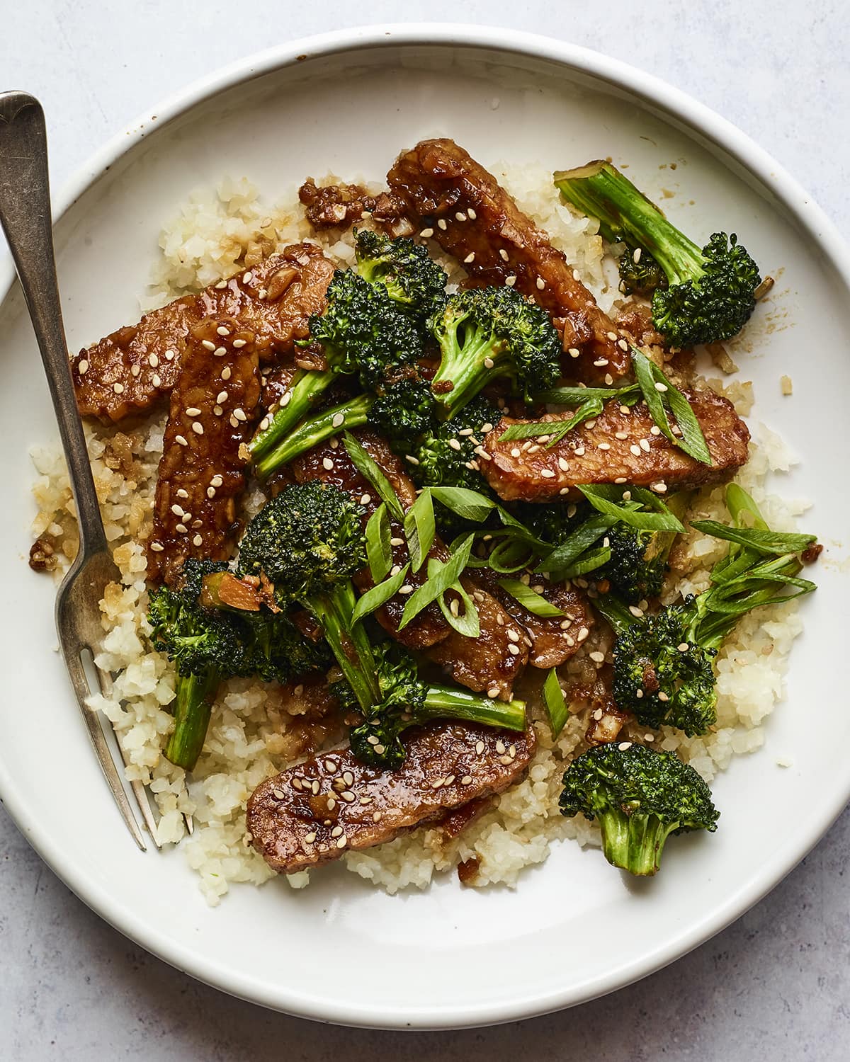 A bowl with rice topped with teriyaki tempeh and broccoli and sesame seeds. A fork is in the bowl.