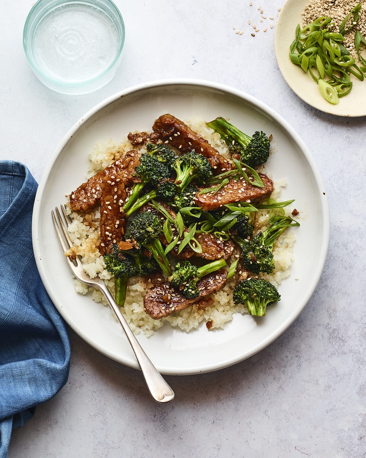 A serving of teriyaki tempeh broccoli served over white rice and garnished with green onion and sesame seeds.