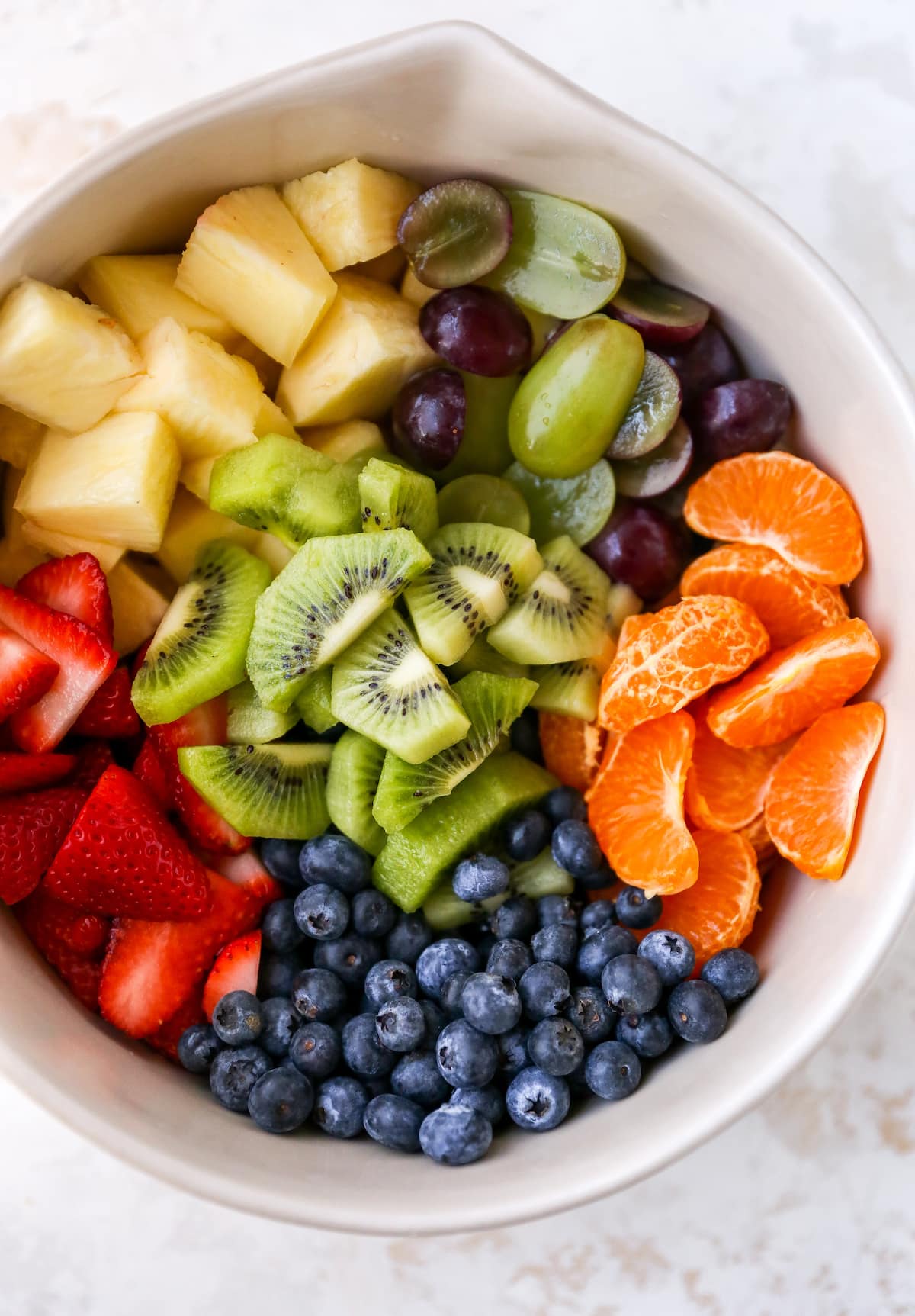 Mixing bowl with pineapple chunks, grapes, clementine segments, kiwi slices, blueberries and strawberries.