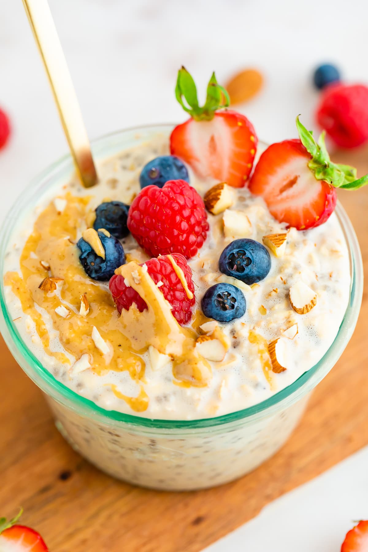 A glass jar of protein overnight oats topped with crushed almonds, fresh strawberry, raspberry, blueberries, and a drizzle of peanut butter.