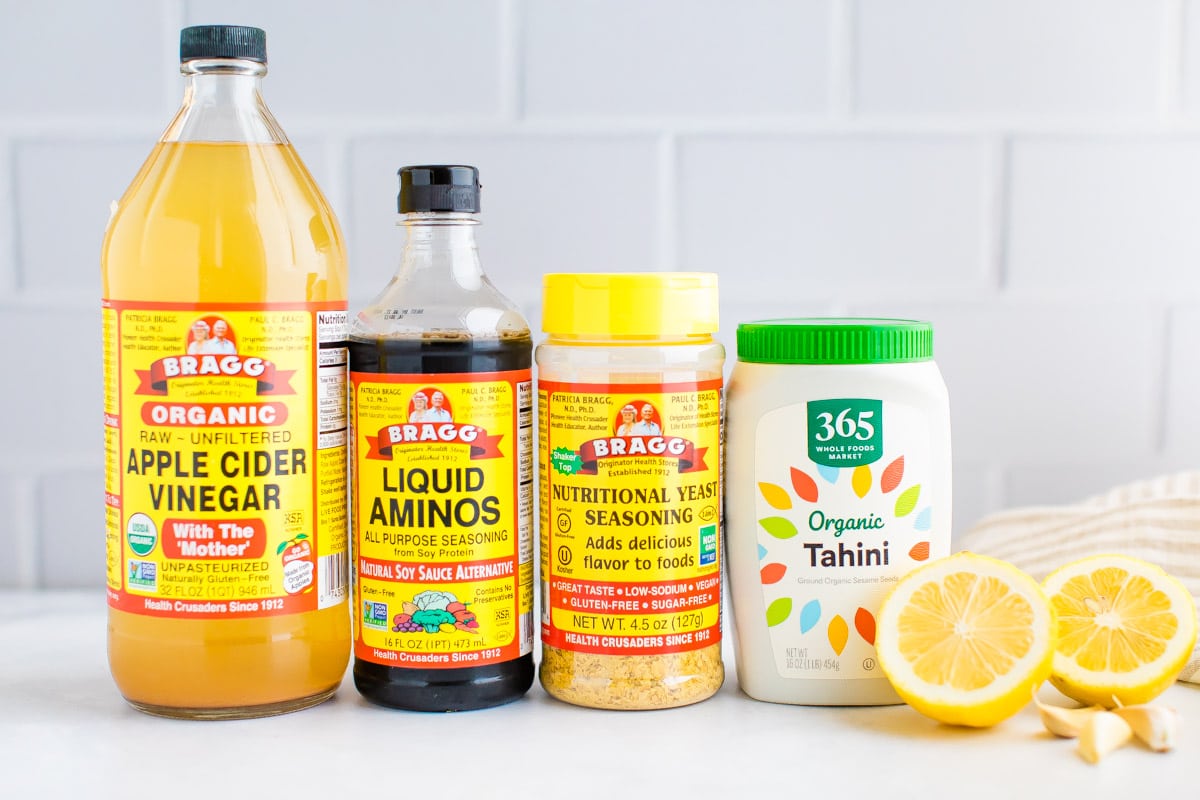Vinegar, liquid aminos, nutritional yeast, tahini, lemons and garlic displayed on counter.
