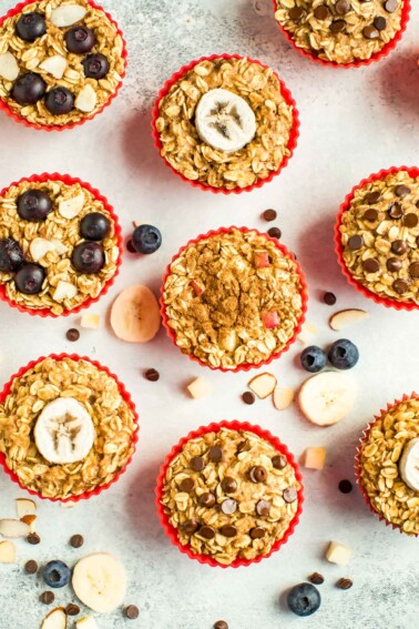 Muffin liners with baked oatmeal cups 4 ways. ingredients are surrounding them on the table like chocolate chips, blueberries, and apple chunks.