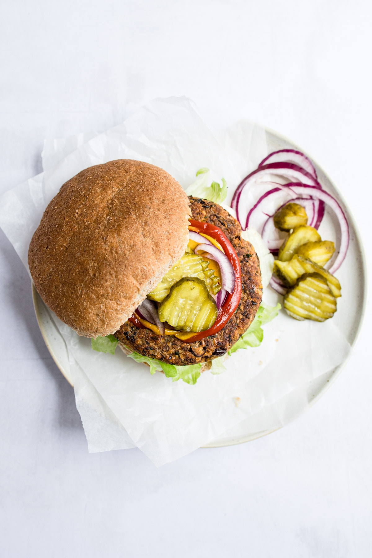 A serving of a black bean burger on a plate and topped with red onion, pickles, and ketchup.