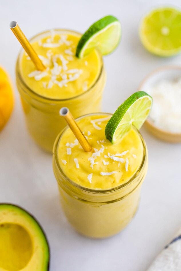 Two glass jars with avocado mango smoothies, topped with shredded coconut, a lime slice on the rim, and a metal straw.