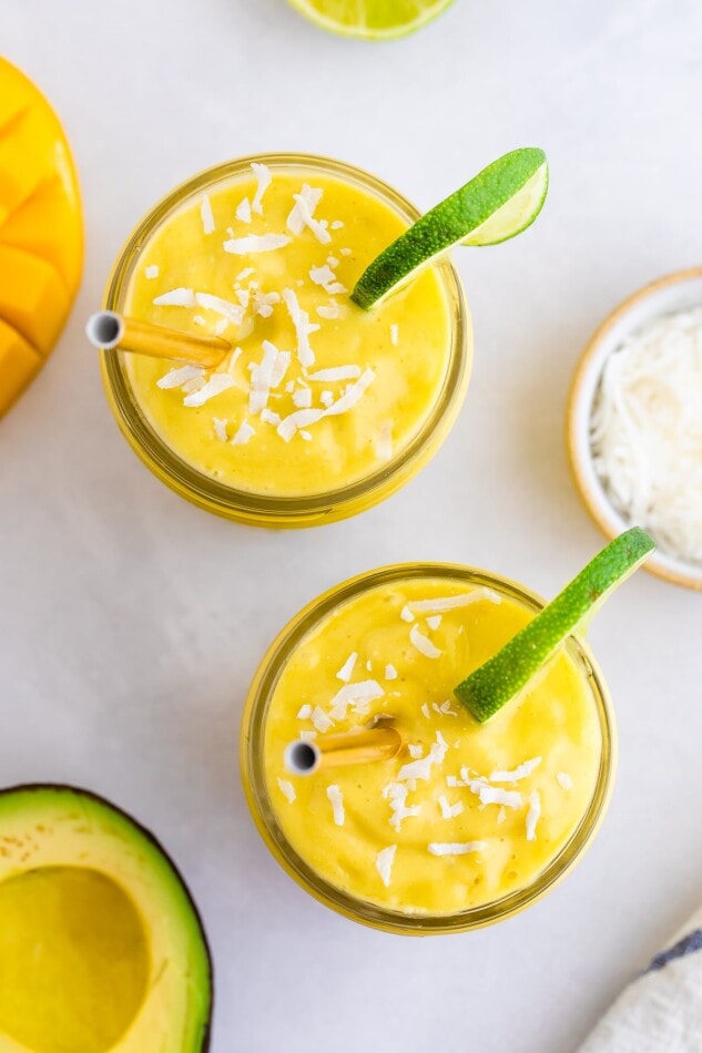 Two glass jars with avocado mango smoothies. They both have metal straws and are garnished with shredded coconut and a lime wedge.