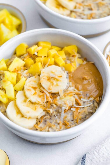 A serving of overnight oats topped with shredded coconut, banana slices, mango, pineapple, and nut butter.