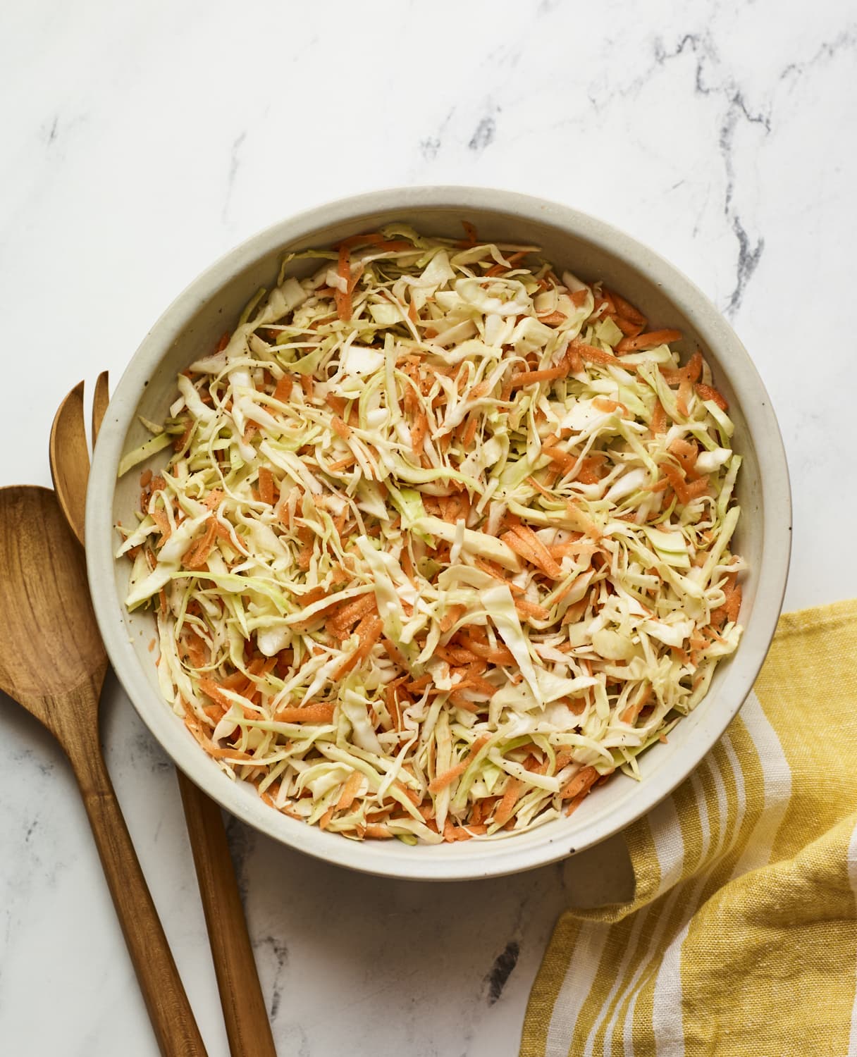 Serving bowl of vinegar slaw. Wood serving spoons and a dish cloth are next to the bowl.