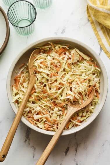 A large serving bowl of vinegar coleslaw with wood serving spoons.