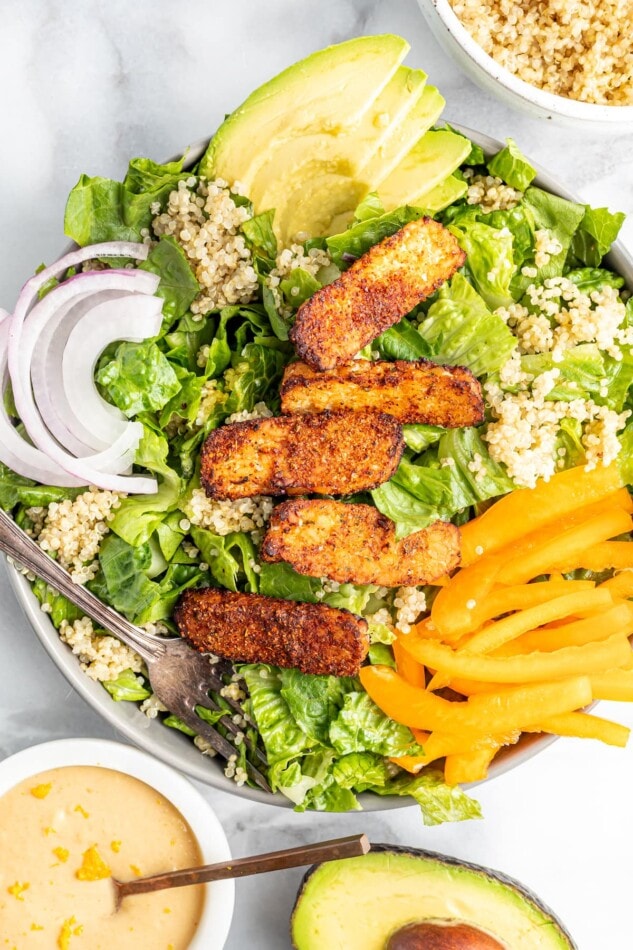 Plant-powered salad with blackened tempeh over fresh romaine with quinoa, red onion, bell pepper, avocado and a creamy orange tahini dressing. Salad topped with blackened tempeh, peppers, onions, avocado and quinoa.