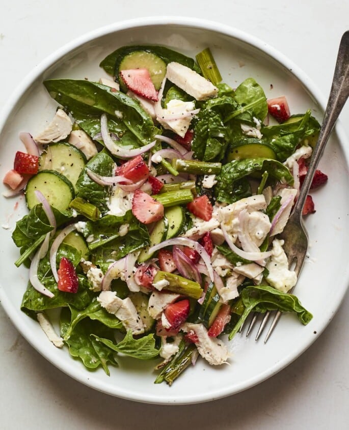 A serving of a spinach salad with shredded chicken, strawberries, cucumber, and red onion on a large plate.