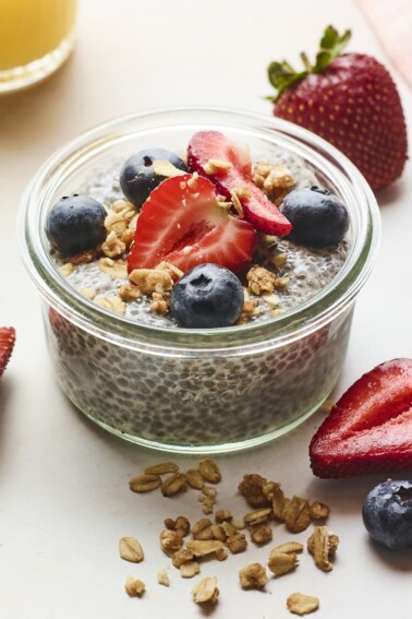 Jar of chia pudding topped with berries and granola. More granola and berries are scattered on the table around the jar.